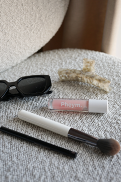 Przezroczysty błyszczyk Pheym. Wonderland na szarej tkaninie. Obok okulary, spinka i pędzel. Efekt 'wet look'.