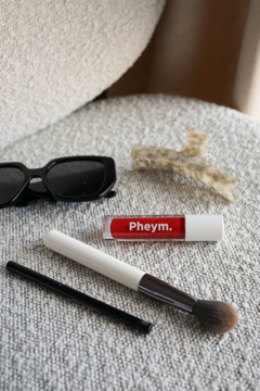 Czerwony błyszczyk Pheym. Cherish, pędzel do makijażu, okulary i spinka na beżowej tkaninie.