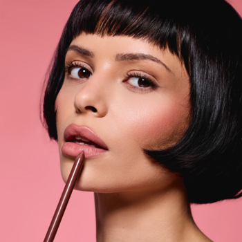 Kobieta aplikuje kremową konturówkę Everybody London Nude Pink. Zbliżenie na usta, różowe tło.