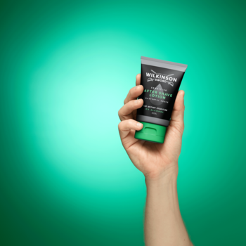 Ręka trzyma nawilżający balsam po goleniu Wilkinson Sword Sensitive w szarej tubce z zieloną nakrętką na zielonym tle.