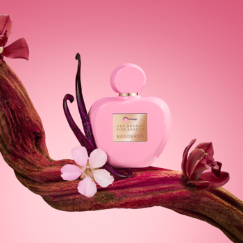 Elegancka, różowa butelka perfum Banderas Her Secret na zakręconej gałęzi z wanilią i kwiatami.
