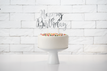 Srebrny, lustrzany topper na tort "Happy Birthday" na białym torcie z posypką na tle białej ściany.