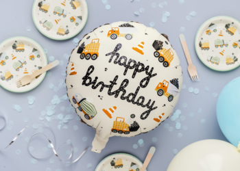 Okrągły biały balon urodzinowy z napisem "Happy Birthday" i grafiką pojazdów budowlanych, otoczony talerzykami i konfetti na jasnoniebieskim tle.