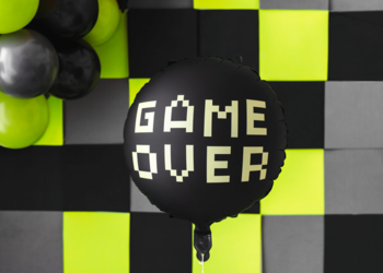 Okrągły czarny balon foliowy z beżowym pikselowym napisem "GAME OVER", na tle zielono-czarnej szachownicy.