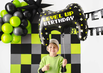 Czarny balon foliowy Level Up Birthday w kształcie gamepada, trzymany przez chłopca na tle gamingowej dekoracji.
