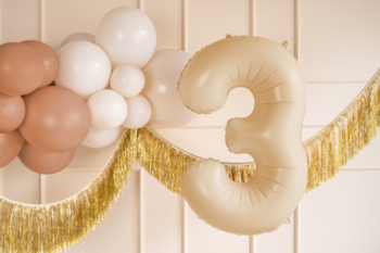 Beżowy balon foliowy w kształcie cyfry 3 na białej ścianie, z pastelowymi balonami i złotą girlandą z frędzlami.