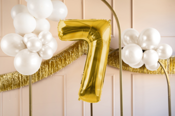 Złoty balon foliowy w kształcie cyfry 7, satynowy, z klastrami białych balonów na jasnym tle.
