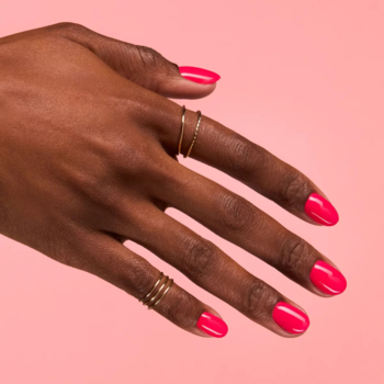 Zbliżenie dłoni z neonowym, malinowym lakierem hybrydowym Semilac Neon Ruby, złote pierścionki, różowe tło.