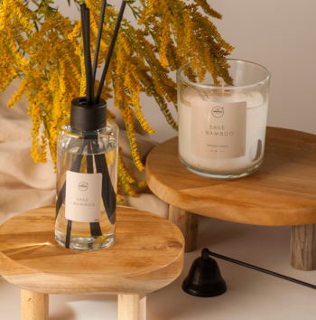 Aroma Home Sage & Bamboo: świeca i dyfuzor z patyczkami na drewnianych podstawkach, z boku z żółtymi kwiatami.