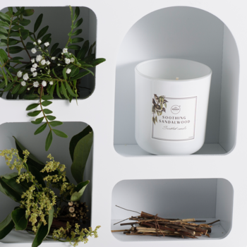 Biała świeca Aroma Home "Soothing Sandalwood" w szklanym pojemniku, w białej niszy, otoczona gałązkami i drewnem.