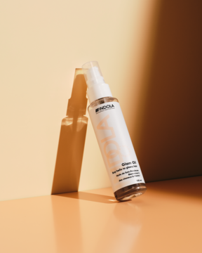 Indola Glam Oil, nabłyszczający olejek do włosów w przezroczystej butelce z atomizerem, oparty o ścianę.
