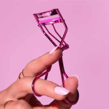 Różowa zalotka Real Techniques Miracle Eyelash Curler, trzymana w dłoni. Podkręca rzęsy, różowe tło.
