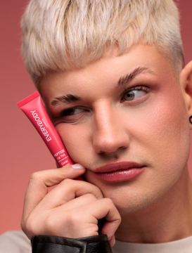 Koralowy tint Everybody London Peptide Lip Tint w małej różowej tubce, trzymany przez modela przy twarzy.