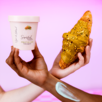 Fluff Superfood jogurt do ciała pistacjowy w kremowym pojemniku. Dwie dłonie, jedna trzyma jogurt, druga croissant pistacjowy. Krem na ramieniu.
