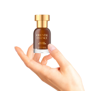Dłoń trzyma brązową butelkę perfum Oriental Essence ze złotą zakrętką, na etykiecie "Golden Magnolia", białe tło.