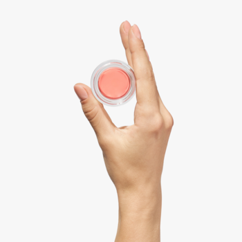 Ręka trzyma pielęgnujący róż Radiant Peach w małym, okrągłym słoiczku. Kremowa formuła do policzków.