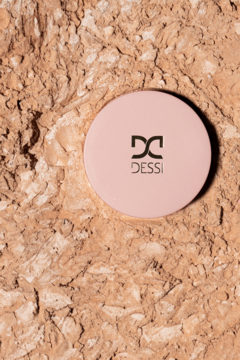 Puder rozświetlający Dessi Glow Out Powder, różowe okrągłe opakowanie z logo, na beżowej skale, widok z góry.