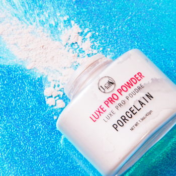 Sypki puder matujący J.Cat Beauty Luxe Pro Powder Porcelain, biały słoiczek na lśniącym niebieskim tle z rozsypanym pudrem.