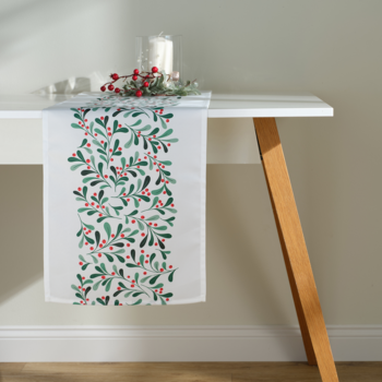 Biały bieżnik na stół z motywem jemioły i czerwonych jagód, na drewnianym blacie, widok z boku.