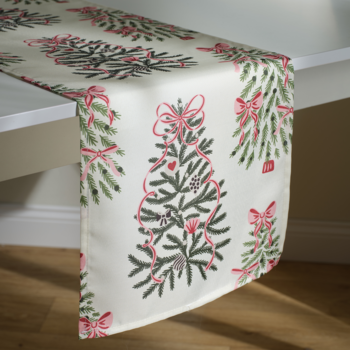 Świąteczny bieżnik stołowy, kremowy z wzorem zielonych choinek, czerwonymi kokardami i bombkami, leży na jasnym stole.