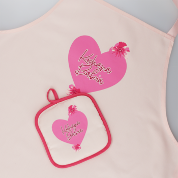 Różowy fartuch i kwadratowa łapka kuchenna z różowymi sercami, złotym napisem 'Kogana Baba' oraz ozdobnymi frędzlami.