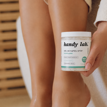 Ziołowa sól do stóp Handy Lab. Patchouli Mood w białym słoiku, trzymana blisko opalonych nóg w łazience.
