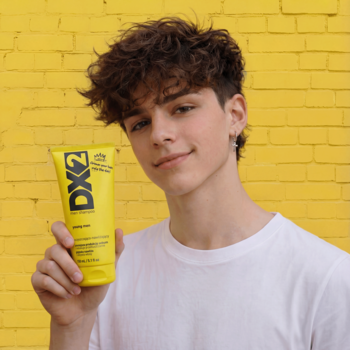 Młody mężczyzna trzyma żółty szampon DX2 Young Men przed żółtą ścianą. Oczyszcza, redukuje sebum.