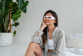 Beautifly SereneEye Pro: kobieta w okularach masujących oczy czerwonym światłem na tle jasnej sypialni.