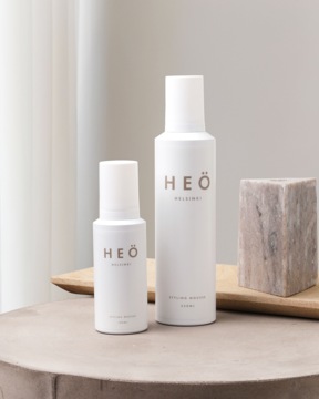 Dwie białe pianki Heö Helsinki Styling Mousse, mała i duża, stoją na drewnianej podstawce z kamiennym blokiem.