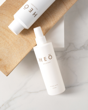 Biała mgiełka nawilżająca Heö Hydra w butelce z atomizerem na marmurze. Obok drugi biały kosmetyk, widok z góry.