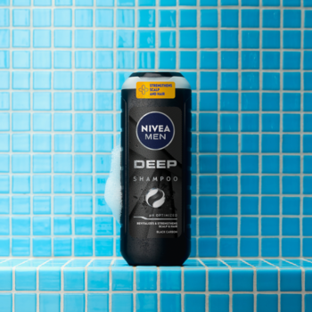 NIVEA MEN Deep Shampoo z czarnym węglem w czarnej butelce, z pianą, na niebieskich kafelkach. Widok z przodu.