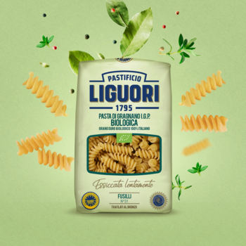 Makaron Fusilli Liguori Integrale BIO w jasnozielonej torbie, otoczony rozsypanym makaronem i świeżymi ziołami. Frontalny widok.