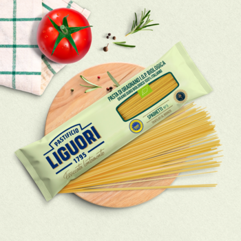 Zielone opakowanie Makaronu Spaghetti Liguori Integrale BIO i nitki na desce z pomidorem i ziołami, widok z góry.