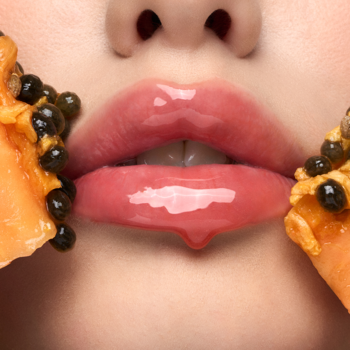 Zbliżenie na błyszczące usta z kroplą olejku, otoczone kawałkami papai. Olejek do ust Exotic Papaya.