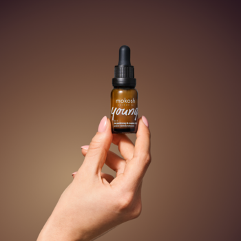 Kobieca dłoń trzyma serum oleiq. young w ciemnobrązowej butelce z pipetą. Na etykiecie napis 'young', brązowe tło.