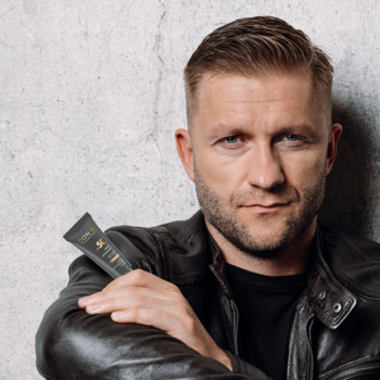 Mężczyzna w skórzanej kurtce prezentuje ciemną tubkę ochronnego kremu do twarzy z filtrem UV, na tle szarej ściany.