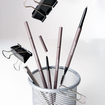 Kredki do brwi Brow Definer Medium Brown z beżową oprawą i szczoteczką, w metalowym kubku, otoczone latającymi spinaczami.