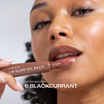 Zbliżenie na kobietę, która nakłada Lumene Lip Glow Oil Balm Blackcurrant na lśniące, śliwkowe usta.