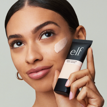Kobieta z próbką e.l.f. Liquid Poreless Putty Primer na policzku, trzymająca czarną tubkę płynnej bazy zmniejszającej pory.