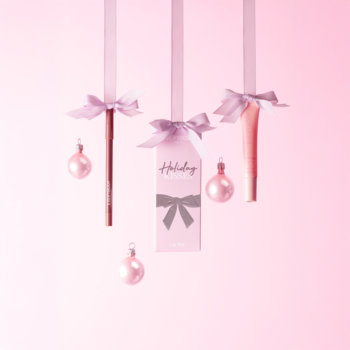 Wiszący zestaw Holiday Kisses: różowa konturówka, balsam do ust i tag prezentowy z bombkami na różowym tle.