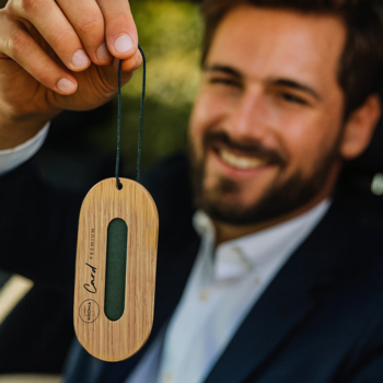 Drewniana owalna zawieszka zapachowa CARD PREMIUM z ciemnozielonym wkładem, trzymana przez uśmiechniętego mężczyznę.