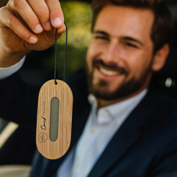 Drewniana owalna zawieszka zapachowa Card Premium, z celulozą w korpusie, trzymana przez mężczyznę w aucie.