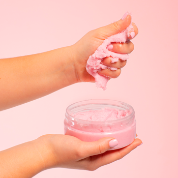 Ręce nabierające różowy cukrowy peeling do ciała Organique Bloom Essence z otwartego słoika.