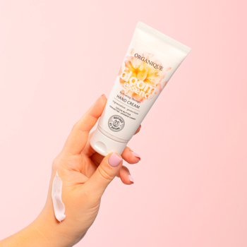 Biały krem do rąk Organique Bloom Essence w tubce, wyciśnięty na kobiecą dłoń na różowym tle.