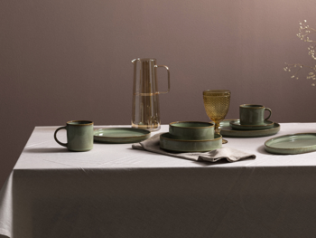 Zestaw naczyń obiadowych w kolorze szałwiowej zieleni z brązowym rantem, na białym obrusie, obok dzbanek.