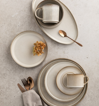 Zestaw kamionkowych naczyń, beżowe talerze, miski i kubki z brązową obwódką, ułożone na szarym blacie, widok z góry.