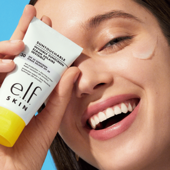 Kobieta uśmiecha się, trzymając e.l.f. SKIN SUNTOUCHABLE, niewidoczny żelowy filtr SPF 30, z próbką na skórze.
