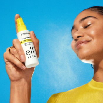Kobieta spryskuje twarz elf SKIN SPF 30 Setting Spray. Widoczna jest delikatna mgiełka z białej butelki z żółtą nakrętką na tle błękitu.