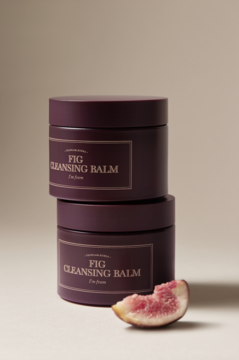 Dwa słoiki I'm From Fig Cleansing Balm w kolorze śliwkowym, ułożone jeden na drugim, obok świeża figa.