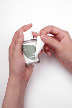 Dłonie odklejają białą etykietę z butelki Isntree Mugwort Calming Powder Wash, ukazując zielony napis.
